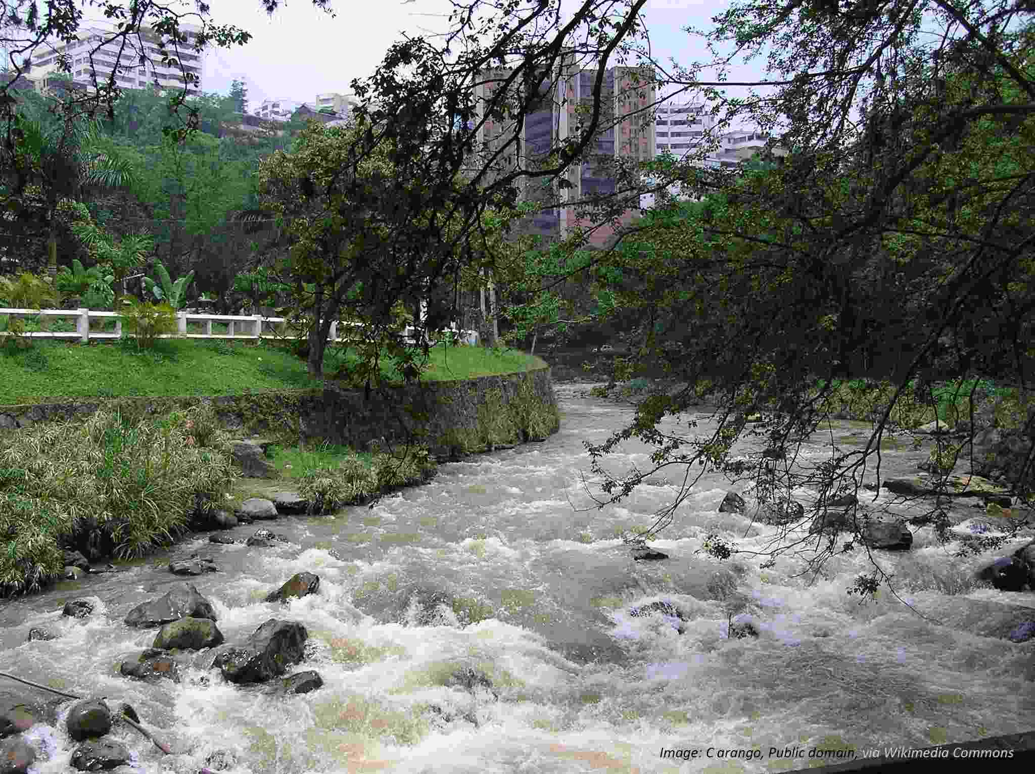 View from a bridge crossing the Cali River, a steep bank is visible in the foreground and the river quickly flowing away from viewpoint. High rise buildings are visible just beyond. 