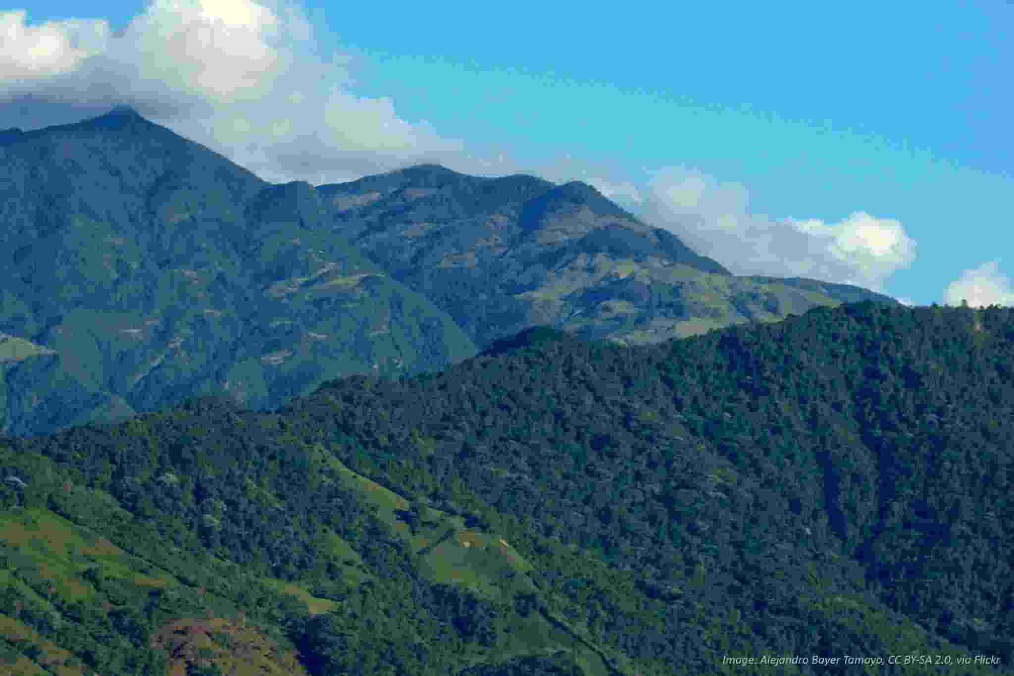 A view across three distinct peaks of the Andes Mountain Range in Colombia, covered in lush green vegetation, against a bright blue sky