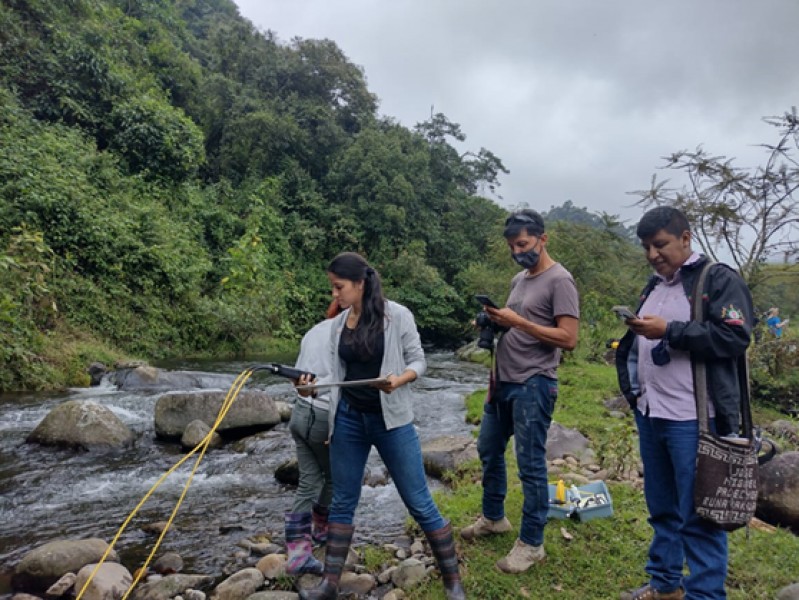 Hub and ASOCAMPO members can be seen taking water quality readings using a multiparameter probe on the bank of the Cauca River, with lush greenery behind