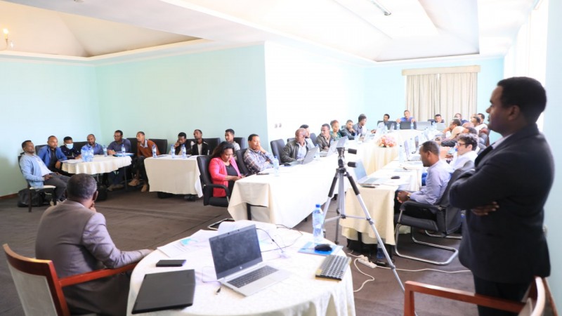 Workshop participants are seated across several tables in a conference room, listening to one of the participants as he speaks. Hub members are leading the session from the front of the room.