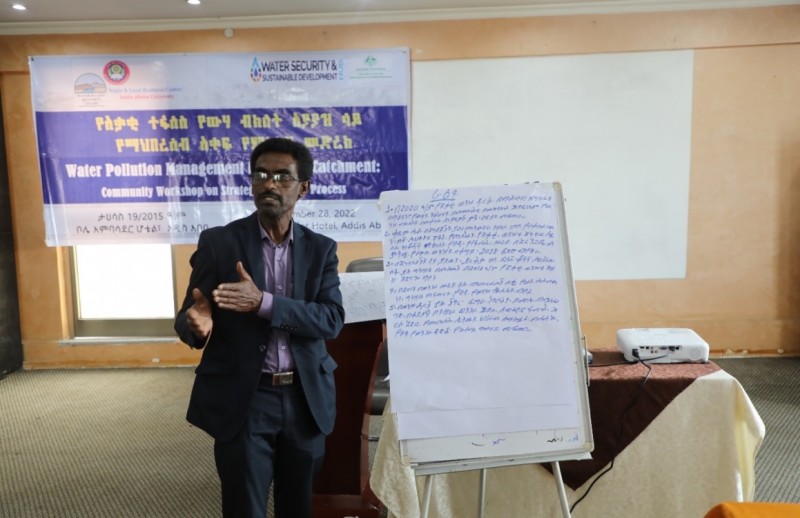 A man stands next to a flipchart as he presents to the audience, with a banner visible behind with the text 'Water Pollution Management in the Akaki Catchment'