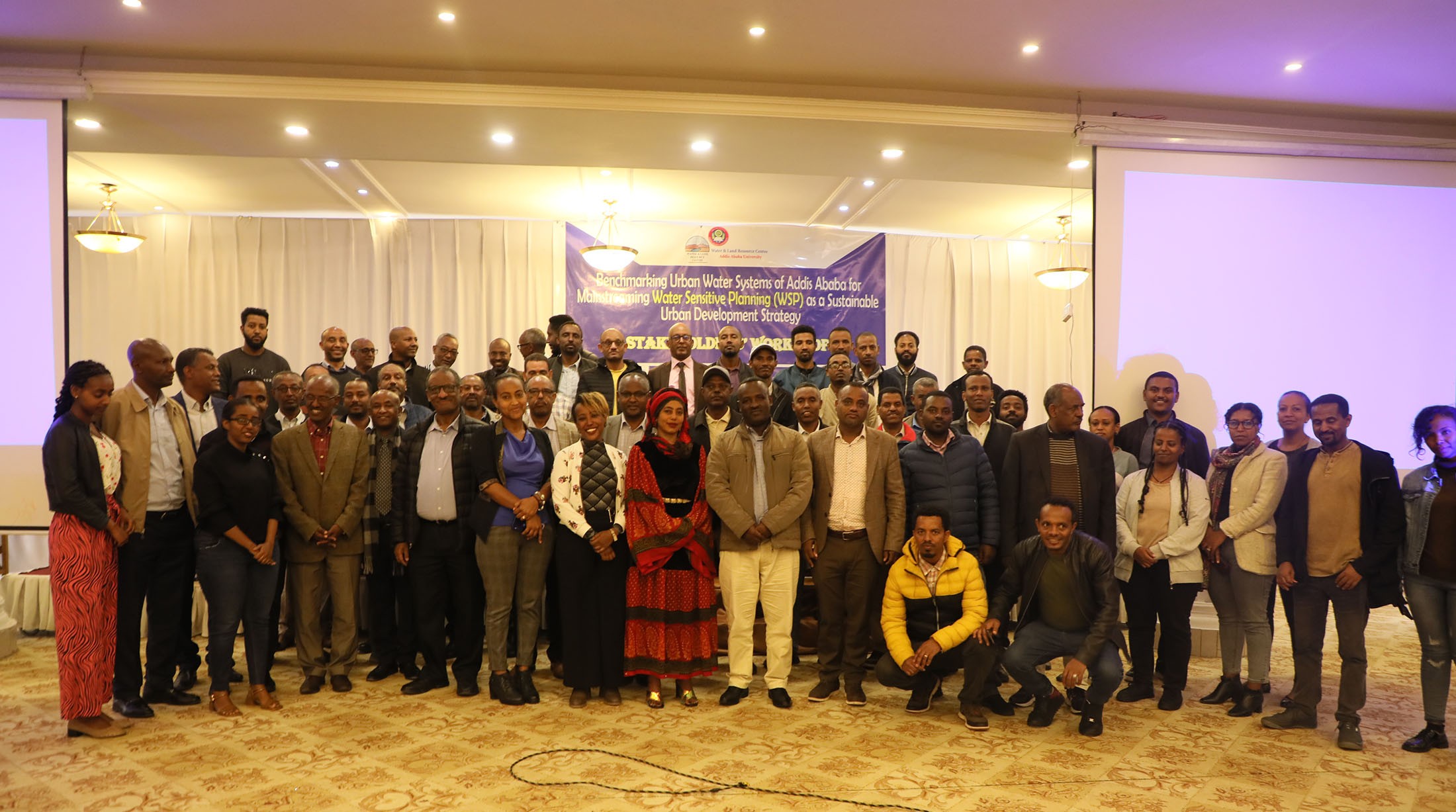 A large group of around 50 people stand and crouch together in several rows, smiling at the camera, with a banner behind with the text 'Benchmarking urban water systems of Addis Ababa'