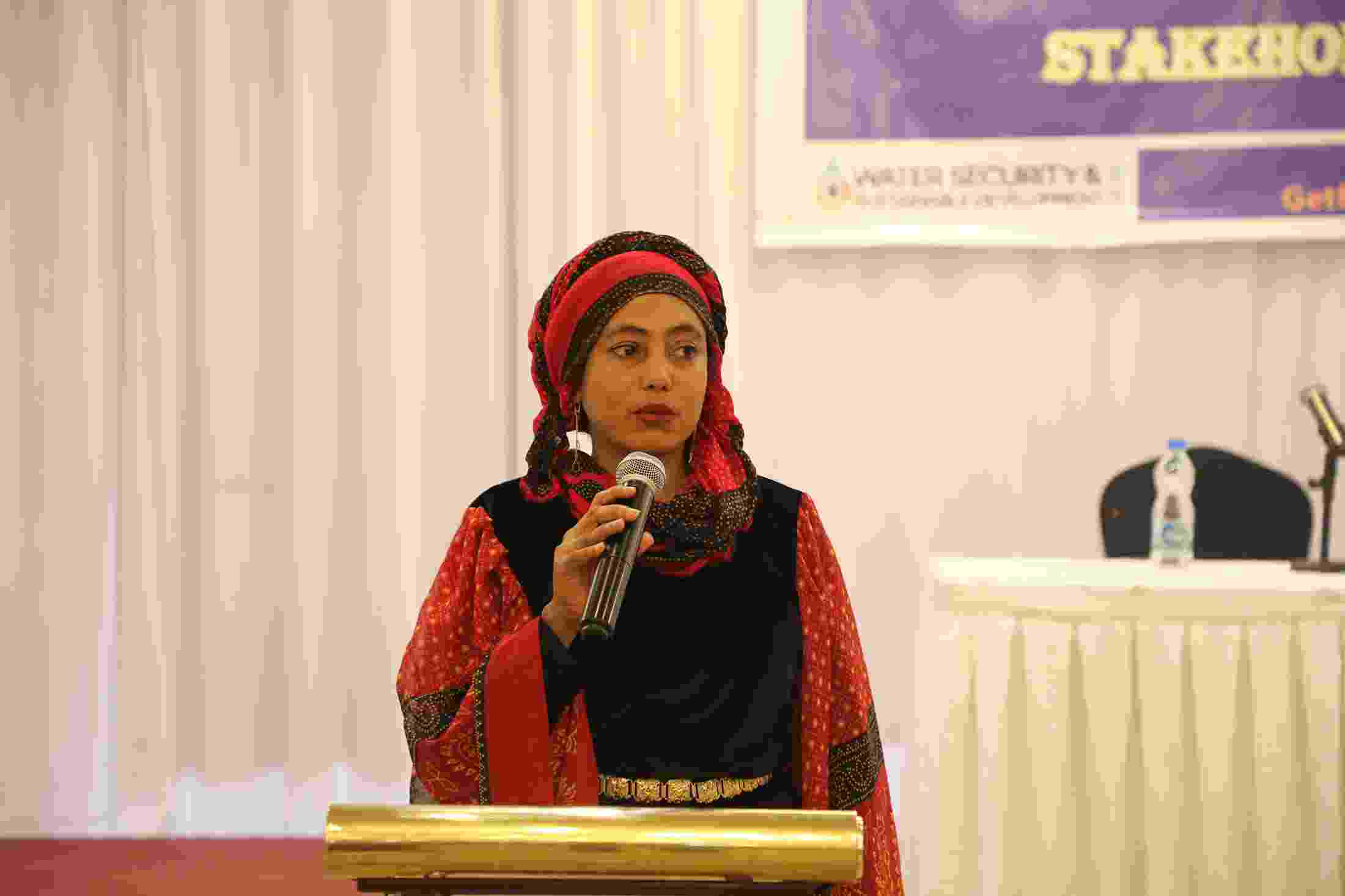 A woman dressed in black and red traditional clothing stands at a lectern and speaks to workshop participants