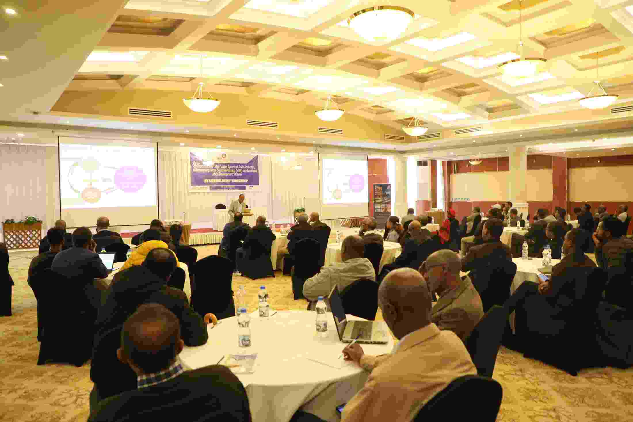 A large room is filled with people sitting together at round tables, facing a male speaker stood at a lectern at the front of the room