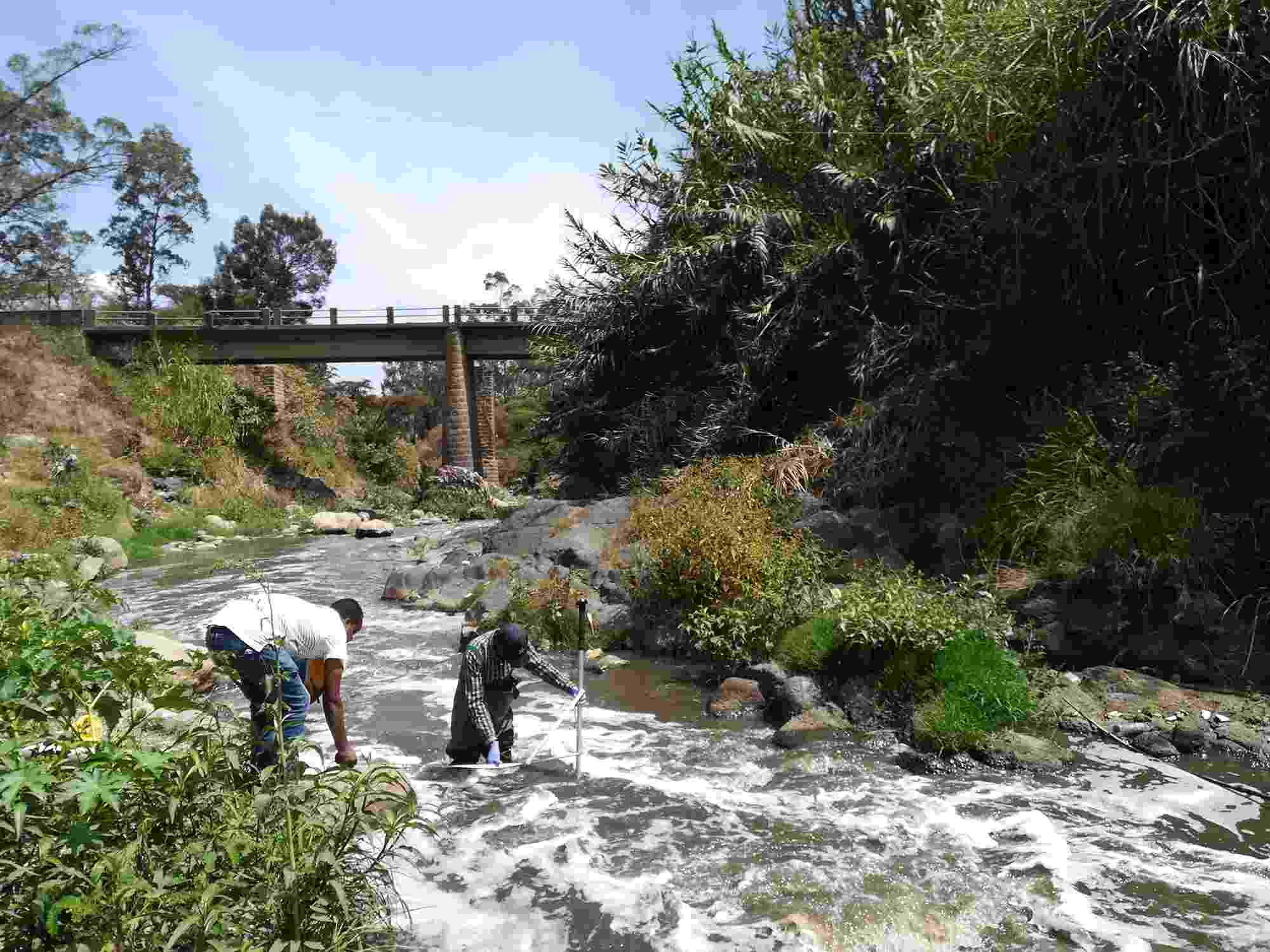 Two Hub colleagues collect water samples from the Akaki River, which flows downstream under a bridge and past rocks and trees
