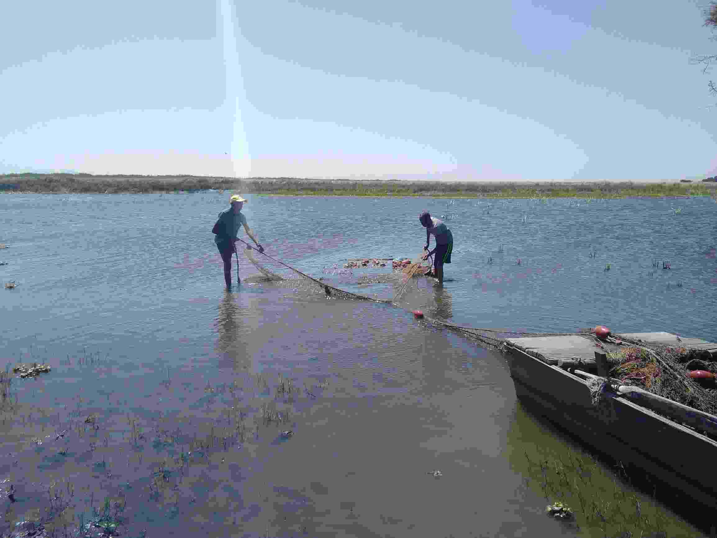 Two men stand ankle-deep in Lake Ziway as they cast fishing nets from their boat on a bright, sunny day