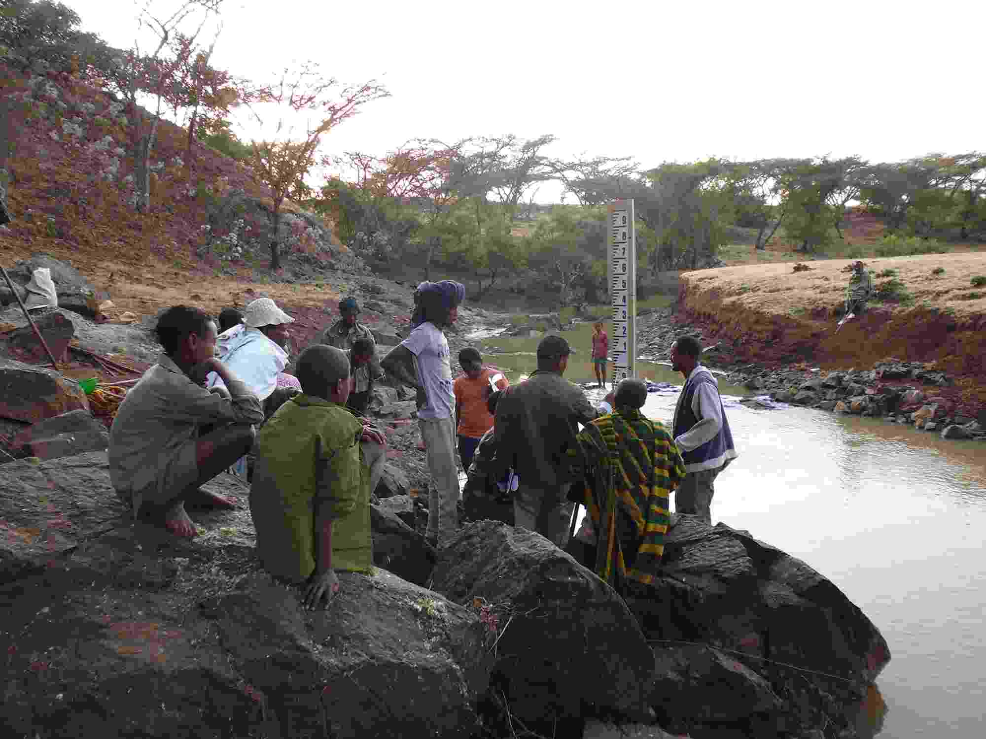 A group of people sit and stand on a rocky riverbank in front of a water depth measurement ruler, whilst listening to a Hub colleage. Trees, shrubbery, and rocks line the riverbanks as it winds away.
