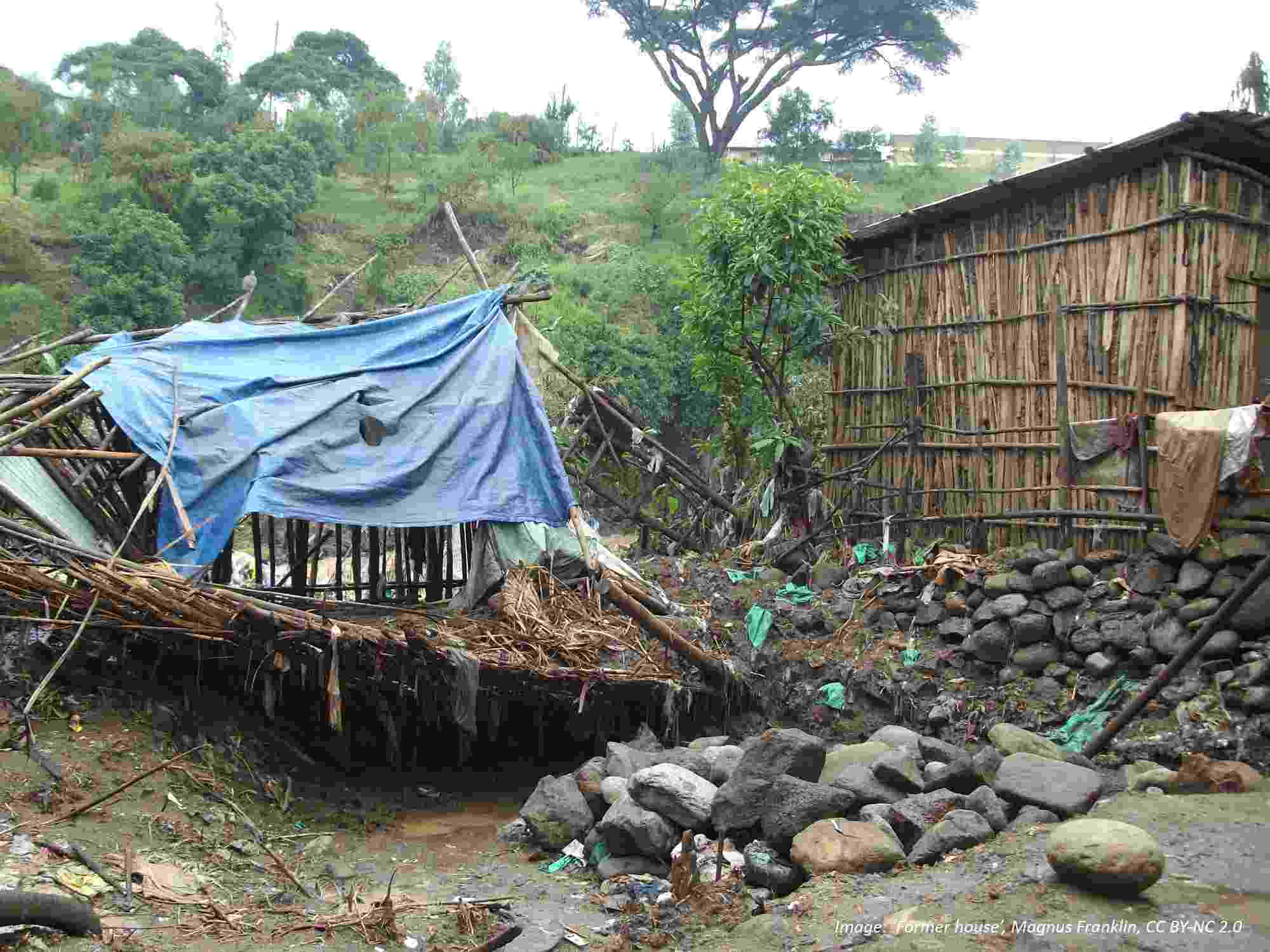 The remnants of a wooden home lie crumpled in a large pile covered by tarpaulin. Flood water has swept away the earth and rock foundation it was built upon. The house next door still stands.
