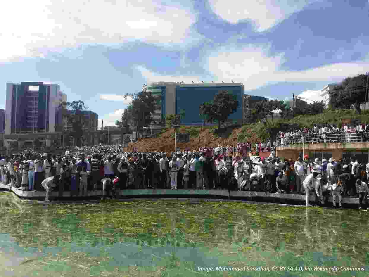 Image shows crowds of people gathered around a pond, celebrating Irreecha with colourful clothing and flowers