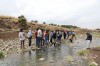 A large group of people stand on the rocky bank of a small river, watching one person as they dip a net into the water to collect a sample thumbnail