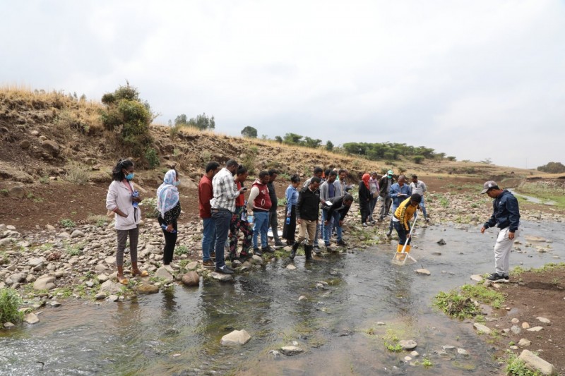 A large group of people stand on the rocky bank of a small river, watching one person as they dip a net into the water to collect a sample