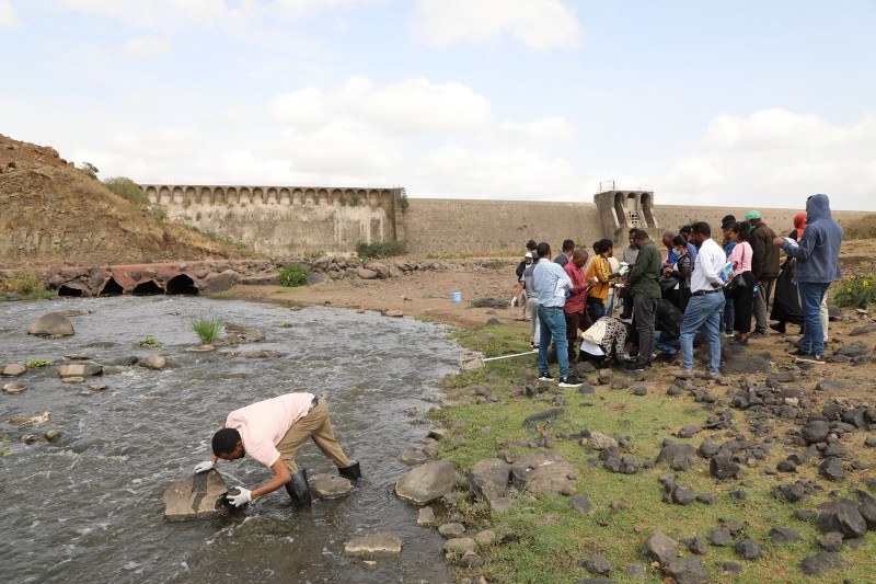 A mixed group of people gather round miniSASS equipment on a river bank, while one man inspects a sampling spot in the river water. A large water dam is visible behind them. 