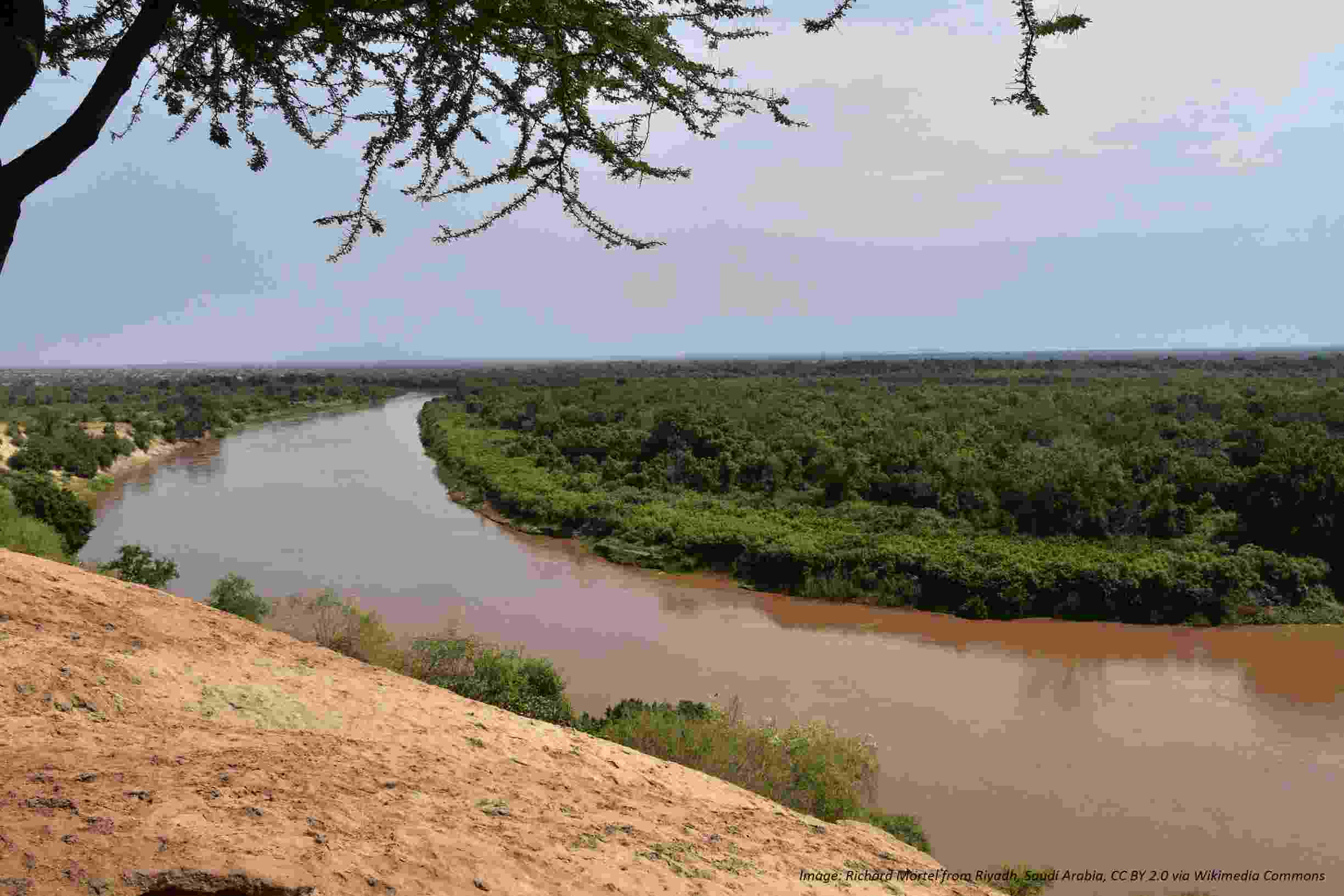 The red water of the Omo River seen from a dusty earth bank as it curves out of sight in the distance, with banks of lush greenery on either side