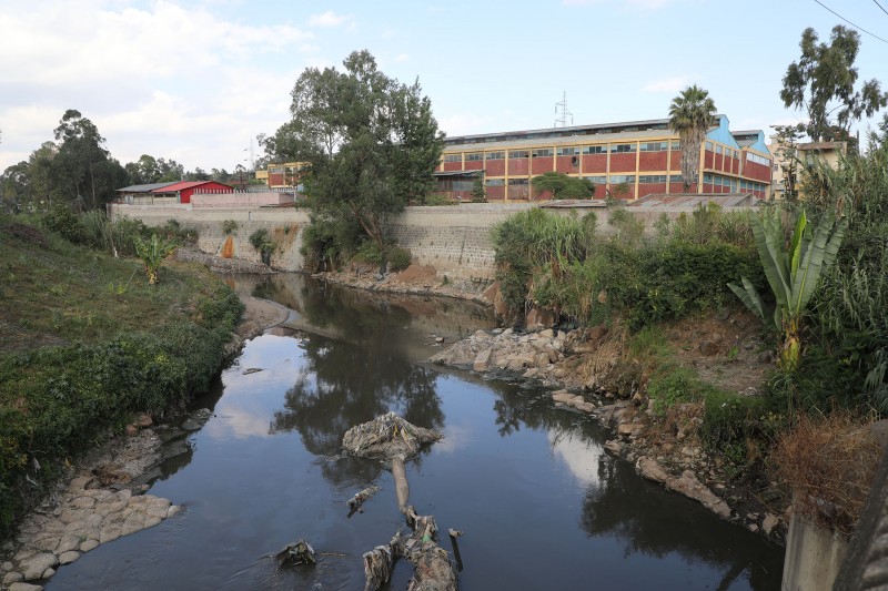 Large factory buildings are located right next to the river water on top of a walled bank, with some bushes, shrubs and palms scattered on either side of the water