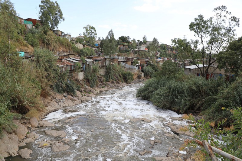 Hundreds of small residential buildings constructed from materials like wood and corrugated metal line and overhang the river, with trees and shrubs scattered among them. The river water looks white and foamy. 