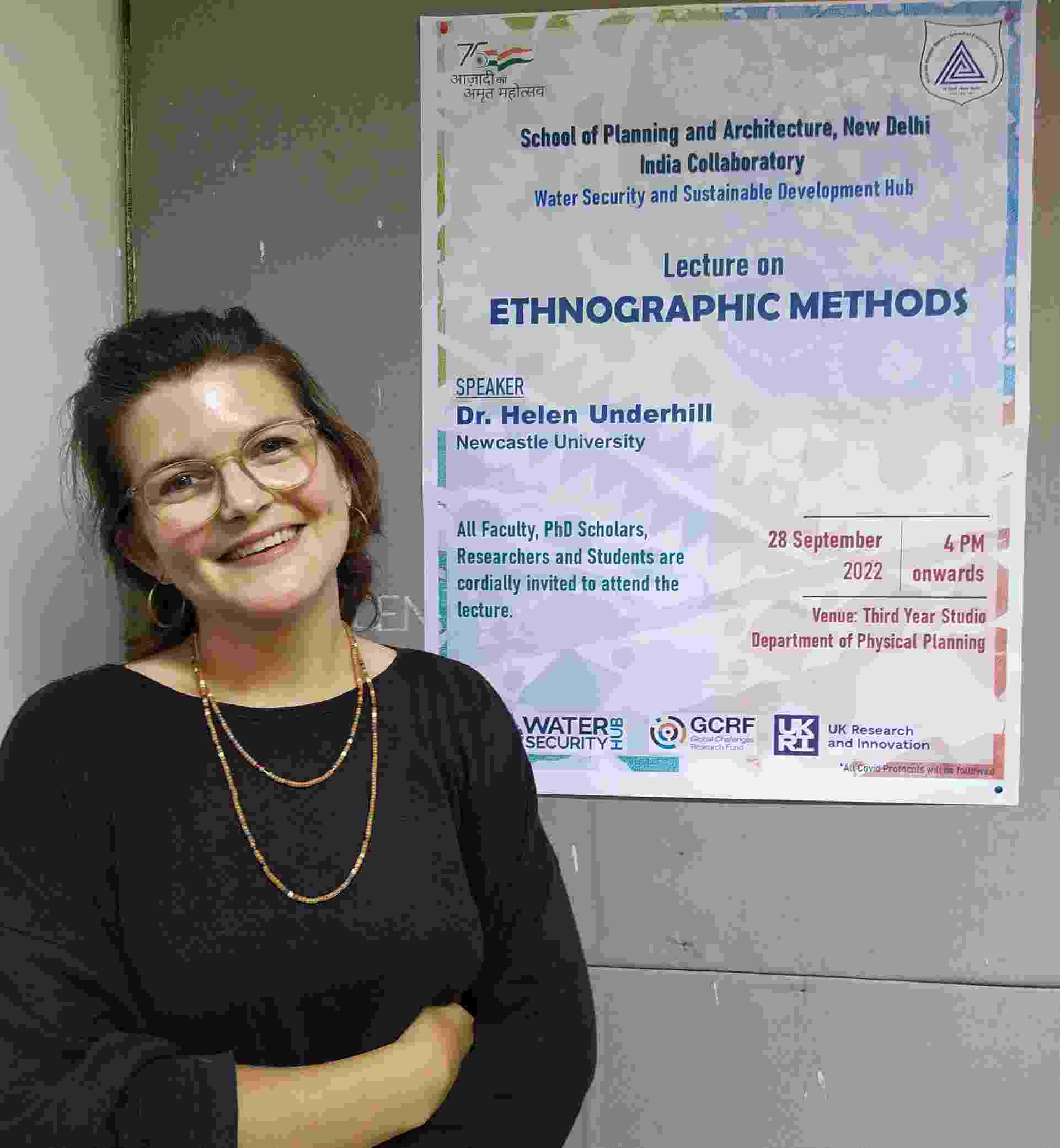 Helen smiles at the camera, stood in front of a poster that reads 'Lecture on Ethnographic Methods'