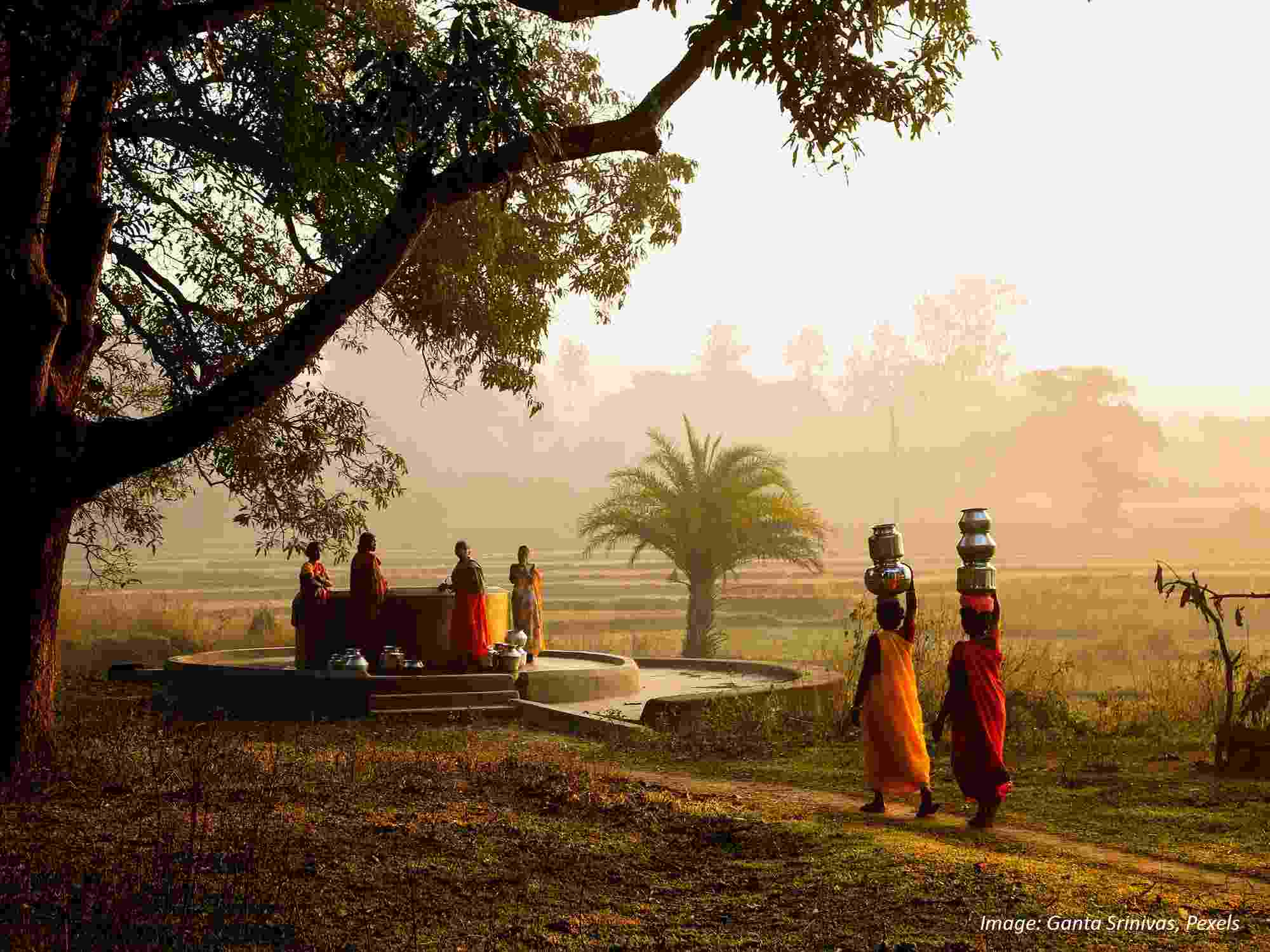 Several women dressed in orange and red on a hazy day, gathered round a groundwater well and carrying water in silver vessels on their heads