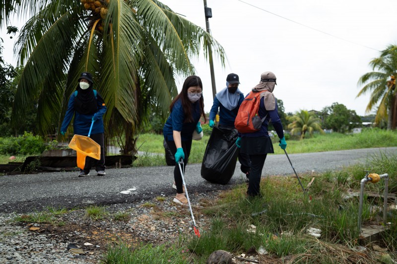 Four female participants in the World Environment Day event collect rubbish and waste using nets, bags, and litter pickers. Two of the participants are stretching forward, using litter pickers to collect items amongst grass and palm trees by a roadside. 