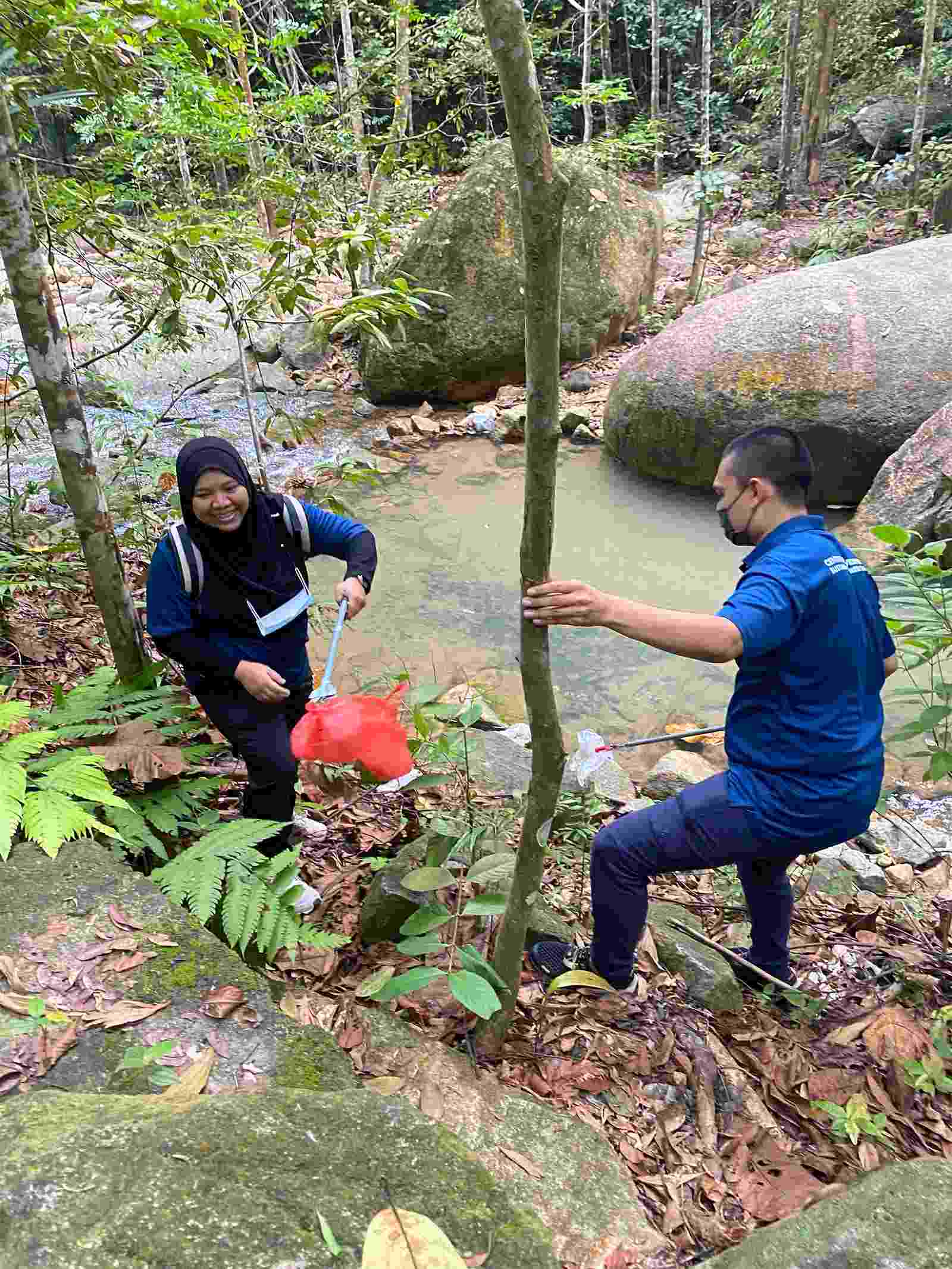 Two Hub researchers clear rubbish from amongst the trees and rocks next to a river