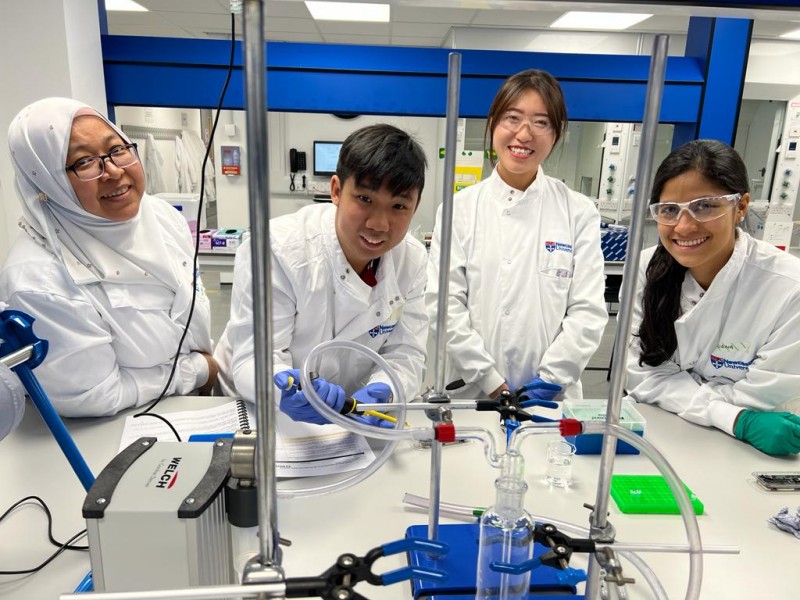Four members of the NUMed team, wearing protective lab clothing, smile at the camera whilst working at a laboratory bench and using water quality analysis tools