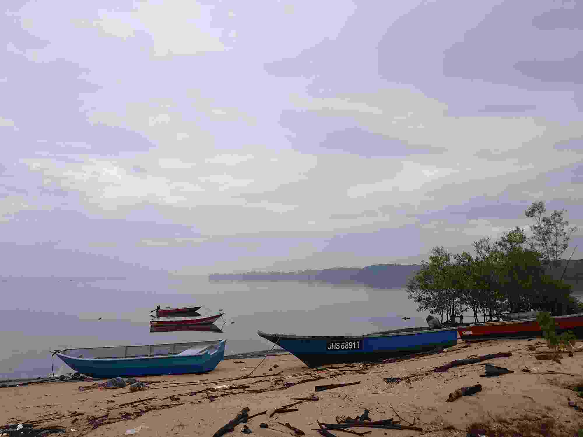 A misty scene on the Johor River in Malaysia, with several boats tethered on the water, and others pulled up onto a sandy bank