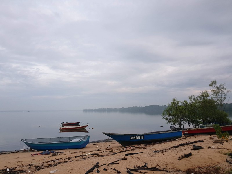 A misty scene on the Johor River in Malaysia, with several boats tethered on the water, and others pulled up onto a sandy bank