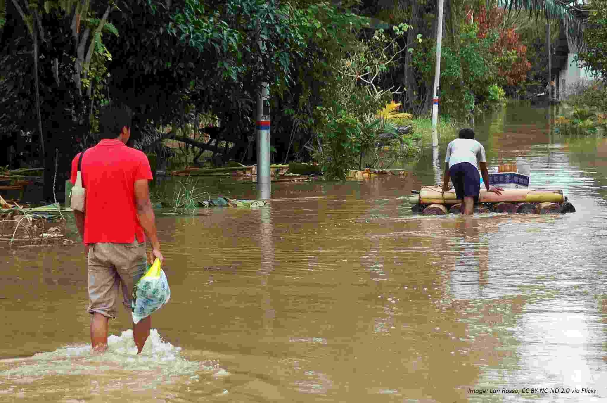 Two men make their way through floodwater that reaches midway up their legs. One is carrying bags, the other is pushing belongings on a homemade raft.