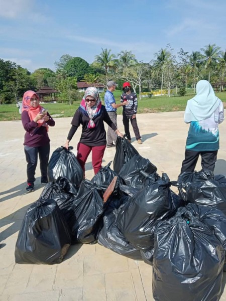 Two women add more full refuse bags to a large pile during the KUDAH event. Other participants and lush greenery and a few buildings are visible behind