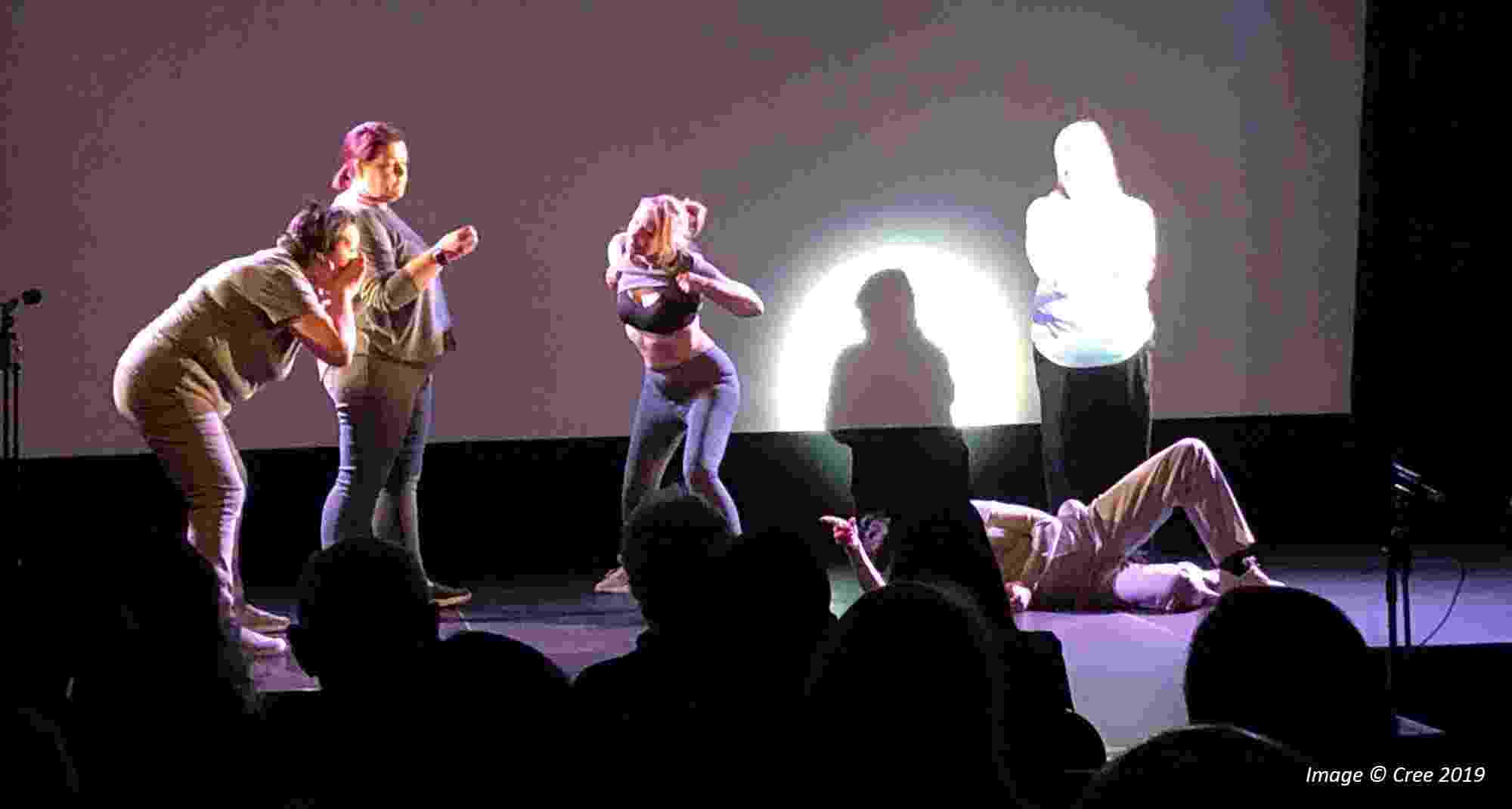 View from within the audience of five people on stage performing under a spotlight