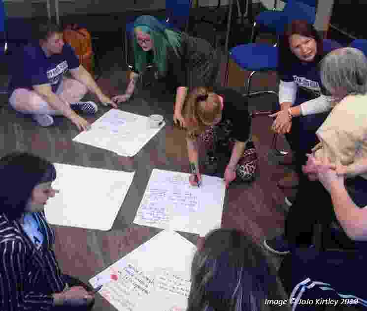 A group of women sit clustered together on chairs and on the floor, holding discussions and working together on pieces of paper scattered between them