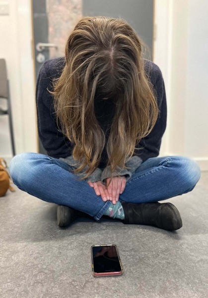 An actress sits cross-legged on the floor, body facing towards the camera with her head bowed close to her chest.