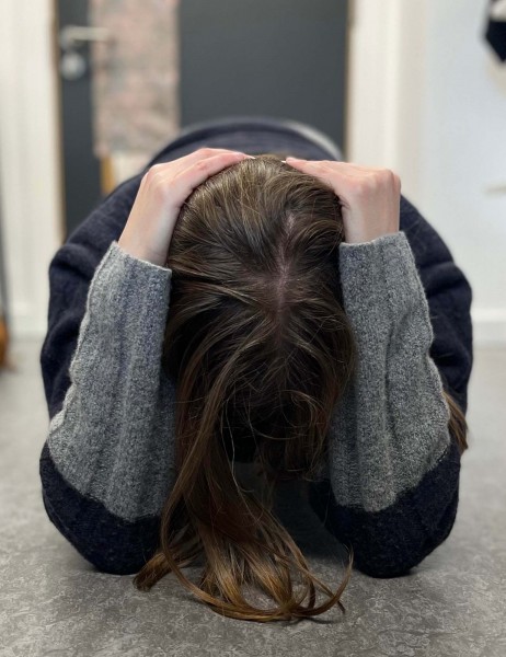 An actress kneels on the floor, cradling her head in her hands with her face turned downwards. 