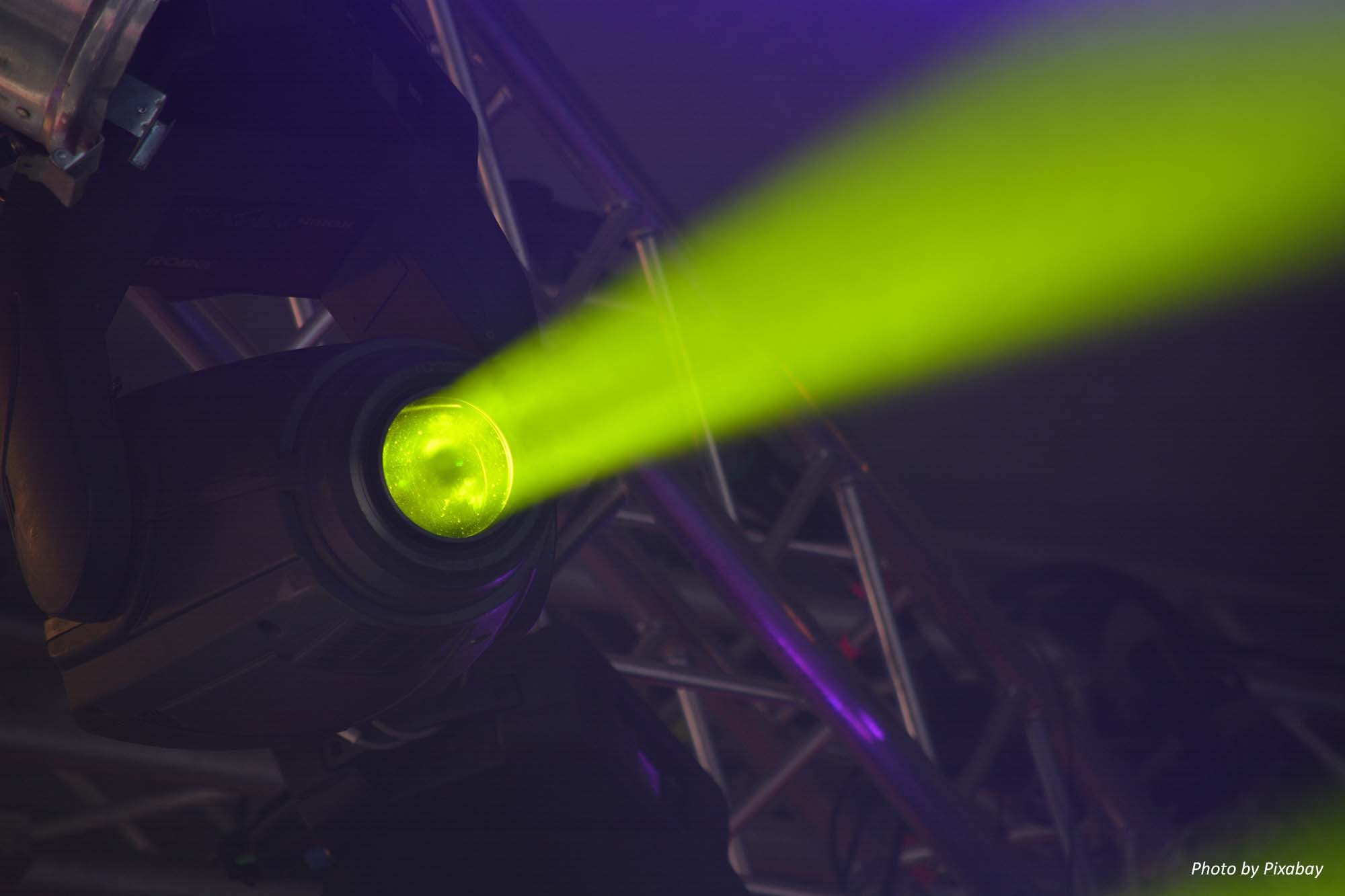 A close up view of a stage light hanging from steel stage rigging, casting a strong yellow beam of light across the darkness