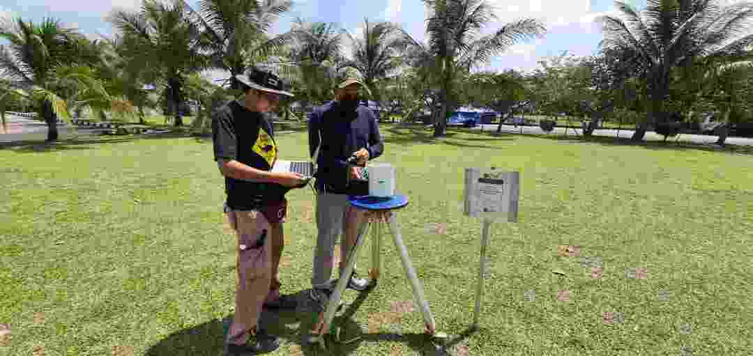 Two hub colleagues collect rainfall data from a sample station located in an open green space, with palm trees behind 